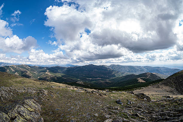 Wide mountain landscape with rocky terrain in the foreground and layered hills stretching to the horizon. Dramatic clouds move across a deep blue sky, creating a natural scene with strong sense.