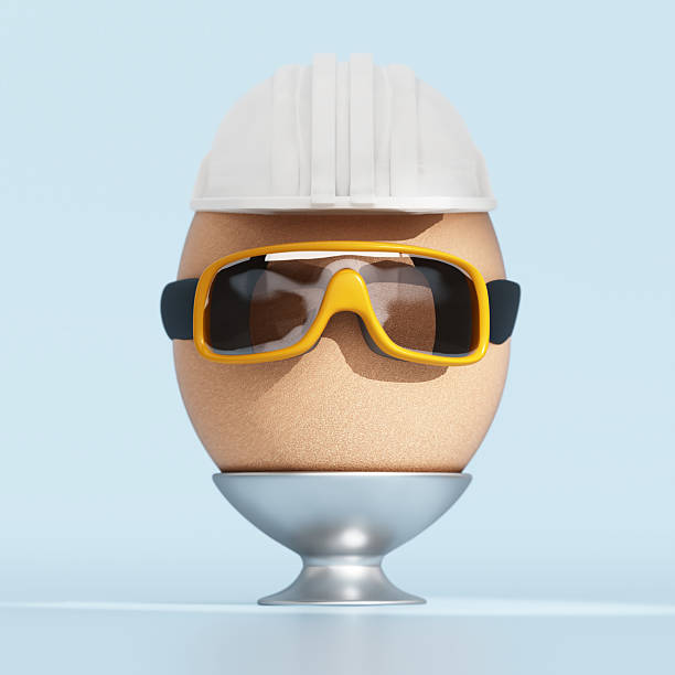 A brown egg wearing a white hard hat and yellow sunglasses is placed in a silver egg holder. The background is a simple light blue color. Protection concept.