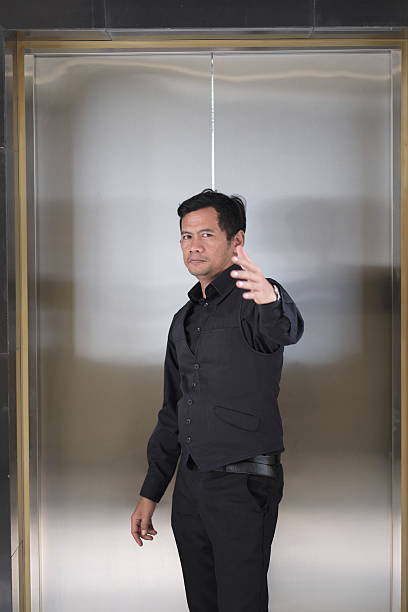 A professional man dressed in black stands confidently in a sleek, modern elevator, looking directly at the camera with a welcoming hand gesture.