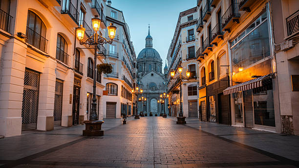 Calle de Alfonso I pedestrian street toward the historic church Basilica of Our Lady of the Pillar  in Zaragoza’s old town at blue hour. Warm streetlights illuminate the buildings, creating a calm and atmospheric urban scene. Zaragoza in Aragón, Spain Europe