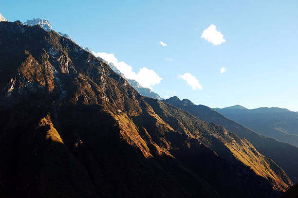 Sunlit mountain range with golden peaks and rocky slopes under a clear blue sky with scattered clouds, showcasing dramatic natural landscape and wilderness scenery.