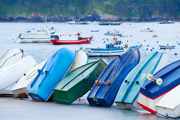Row of blue and green fishing boats leaning upside down on harbor dock. Larger fishing boats moored in the background. Lorbé, A Coruña province, Galicia, Spain.