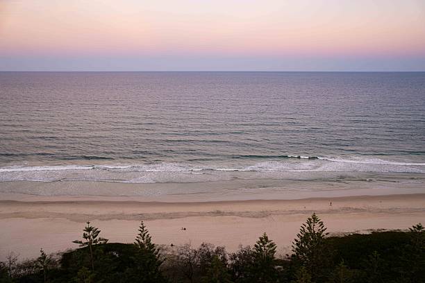 This serene coastal scene captures waves moving gently toward the shoreline as the sun sets, painting the sky in warm yellow tones and creating a calm, atmospheric beach moment