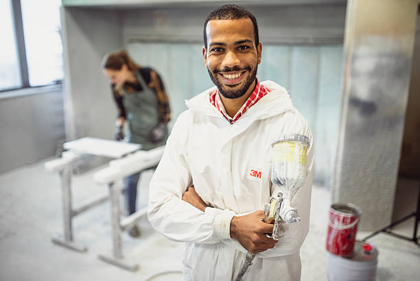 Mid adult multiracial man smiling in workshop while holding paint spray gun, wearing protective coveralls. Workshop setting with tools and equipment in background suggests contentment and professionalism.