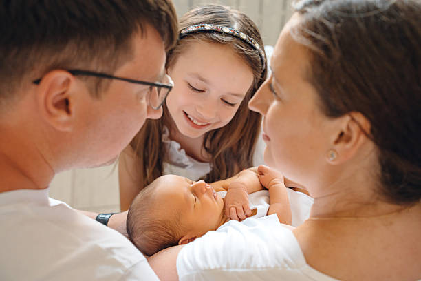 A family is gathered together in a cozy room, with parents holding a newborn baby. A sibling smiles happily while looking at the baby.