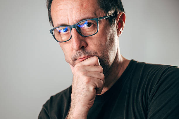 Serious middle-aged man wearing glasses, hand on chin, looking at camera with thoughtful expression, symbolizing intelligence, analysis, and critical thinking. Selective focus.