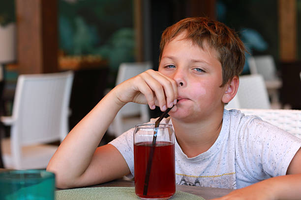Young caucasian boy with blue eyes is drinking red beverage from glass with straw. Kid enjoys refreshment. Child leisure and summer activity concept.