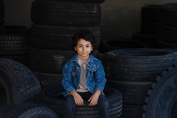Portrait of a smiling little boy sitting on stacked vehicle tires inside an industrial workshop or garage environment.