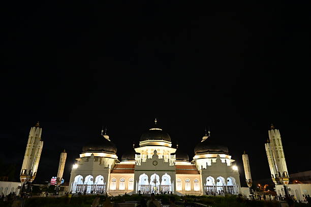 The Baiturrahman Grand Mosque is beautifully illuminated with bright lights at night in Banda Aceh, Indonesia to showcase its grandeur and architectural beauty by lighting up the mosque against the dark night sky, highlighting its majestic features