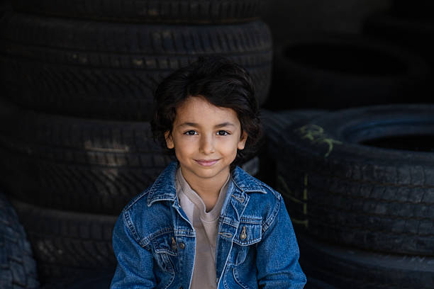 Portrait of a smiling little boy sitting on stacked vehicle tires inside an industrial workshop or garage environment.