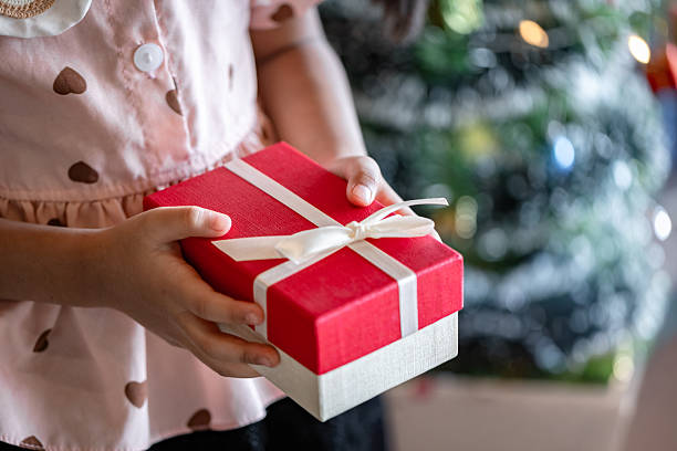 istock Child's Hands Holding a Red and White Christmas Present Against Blurred Festive Tree Background 2252006889