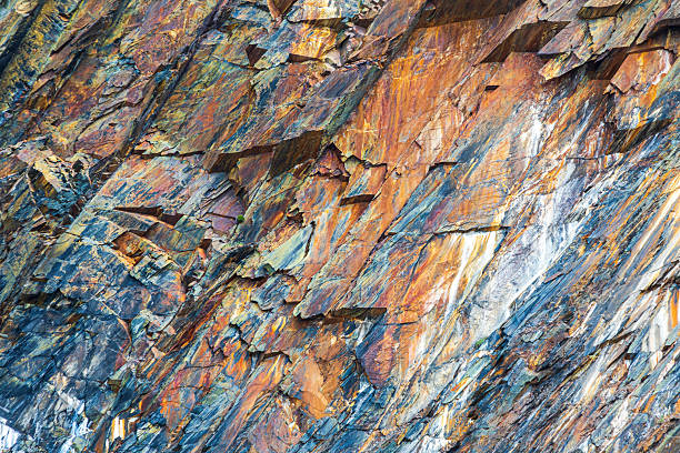 A close-up of a rugged rock wall featuring shattered slabs in blues, oranges, and browns. Natural patterns and mineral veins create an abstract, textured surface inspired by geology and nature.