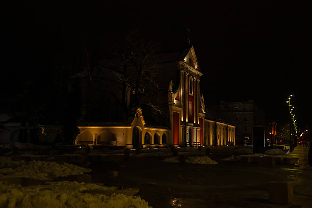 Capuchin monastery of Peter and Paul in Lublin. A building stands illuminated at night. Snow covers the ground, and lights outline the structure. Trees are visible in the distance.