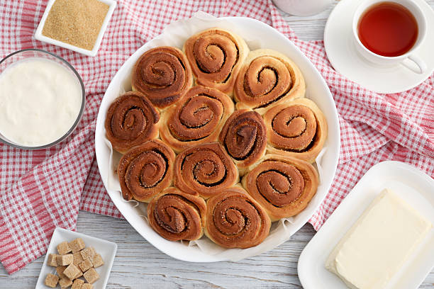 Freshly baked cinnamon rolls, tea and ingredients on light wooden table, flat lay