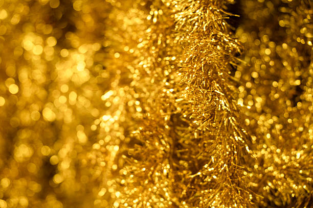 Stunning close-up abstract photograph showcasing luxurious gold and yellow metallic tinsel strands creating dense overlapping textured pattern with brilliant shimmering reflections. Features vibrant golden foil ribbons with warm amber bronze tones capturing light reflections and iridescent effects. Perfect visual content for 2025's biggest retro tinsel comeback trend with maximalist sparkle and nostalgic charm. Ideal for festive backgrounds, Christmas decorations, party invitations, New Year celebrations and creative abstract design projects.
