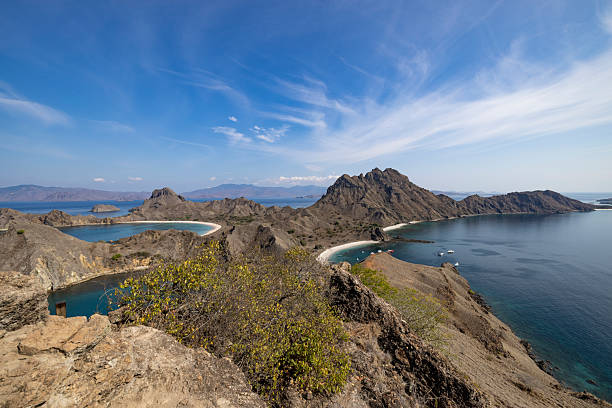 View from the ridge of Padar Island in Komodo National Park, Indonesia, showing multiple turquoise bays, rugged volcanic hills, and dry island terrain under a bright sky with sweeping clouds.