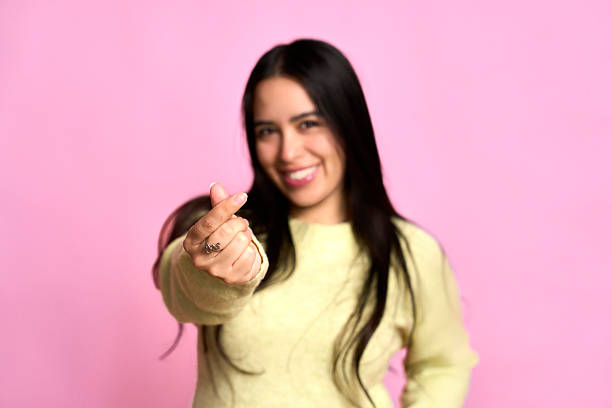 Smiling young adult woman making a finger heart gesture against a pink background