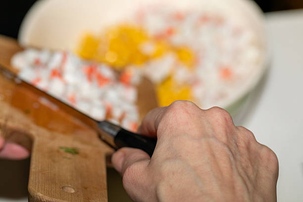 Close-up of a hand adding chopped crab sticks from a wooden board to a salad bowl. Authentic home cooking process with selective focus on the action.