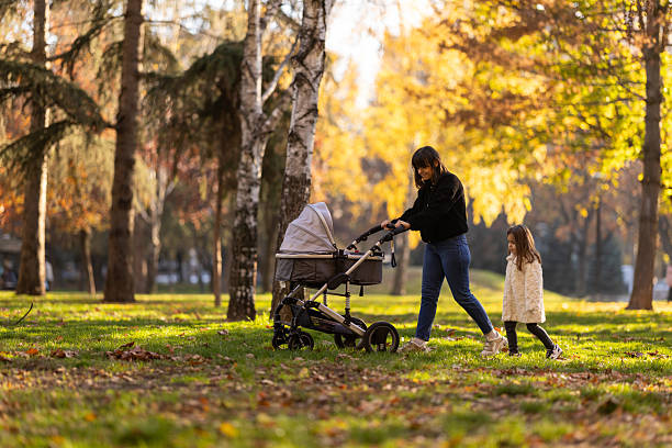 A mother pushes a baby stroller as a little girl walks beside her in a sunlit autumn park. Warm colors, falling leaves, and casual outdoor family time convey care, togetherness, and everyday joy.