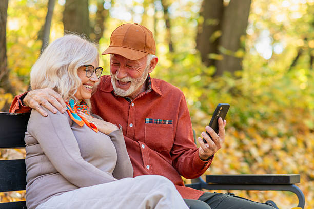 Cheerful senior man and woman relaxing on a park bench, laughing while browsing a smartphone together during a calm autumn day surrounded by colorful fall foliage.