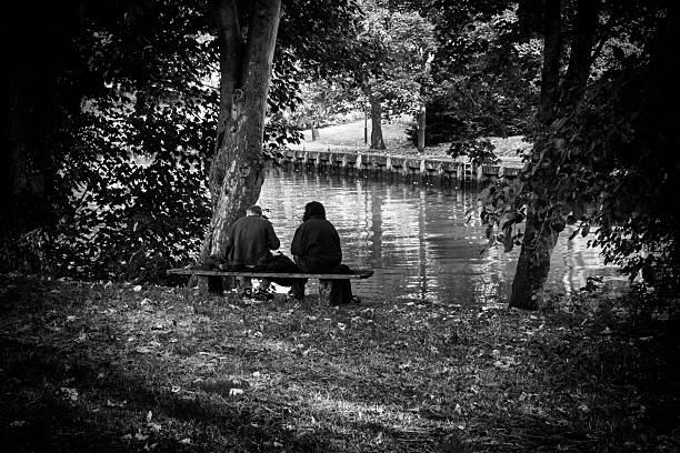 Norwich, Norfolk, United Kingdom: October 14, 2024: Two people sitting on a wooden bench beside the River Wensum in Norwich, Norfolk, Eastern England, watching the river flowing past.