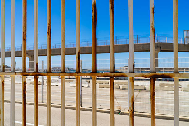 Close-up of a rusted industrial security fence with vertical bars, isolating a blurred elevated road bridge structure against a bright blue sky.