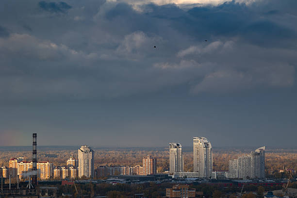 Distant view of Obolon skyscrapers and industrial chimney under dark grey sky. Mood of approaching storm over wide autumn city landscape and Dnipro river horizon. Industrial pollution concept.
