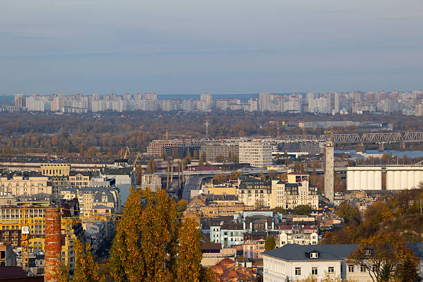 Panoramic view of Kyiv city skyline extending from Podil to Troieshchyna district. Industrial grain elevator and bridges across Dnipro river in autumn. Urban landscape with residential architecture.