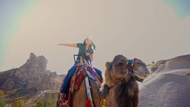 A smiling woman sits on a decorated camel and waves toward the camera while enjoying her time in Cappadocia. The scene highlights adventure tourism and the region’s famous natural formations. Perfect for travel campaigns, social media, and tourism promotions.