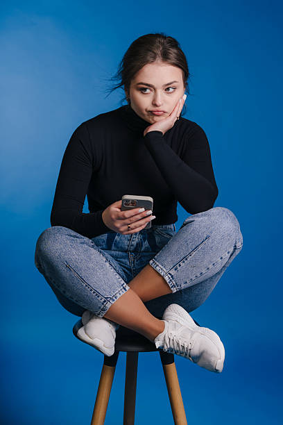 istock Contemplative young woman using smartphone on stool against blue background 2252080217