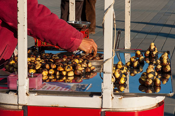 Close-up details of a traditional roasted chestnut vendor stall in Istanbul, Turkey, showing the nuts, a vintage red scale, the seller's hands, and the cart amidst the bustling Taksim square.
