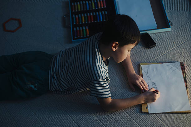 caucasian boy drawing on floor clipboard lamplight overhead, prone position, focused strokes, carpet texture, markers and pencil,
