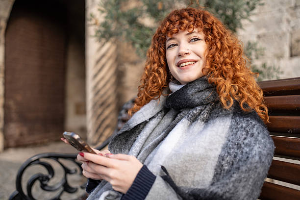 istock Smiling Red-Haired Woman Sitting On a Park Bench Using Smartphone in Cozy Grey Scarf 2252086701