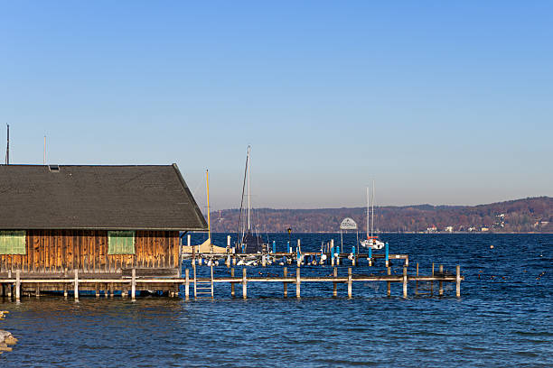 A wooden boat shed with a dark roof sits beside an old dock on Lake Starnberg, south of Munich. The scene opens onto wide blue water, with the masts of docked sailboats rising in the distance. Forested mountain slopes form a scenic backdrop, adding depth and a calm, natural atmosphere to the lakeside setting.