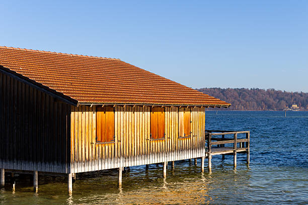 This image displays an old wooden boat shed located on Lake Starnberg, which is located south of the city of Munich. The afternoon sun is shining golden light on the wall of the boat shed, giving it a golden glow, whilst dark blue water fills the midground and a foresty hill sits in the distance.
