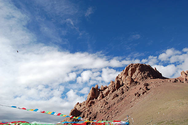 Rocky mountain formation with colorful prayer flags against a blue sky with scattered clouds. Natural landscape featuring rocky terrain and cultural symbols in a serene outdoor setting.