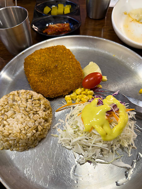 Breaded cutlet served with cooked brown rice, fresh shredded cabbage salad topped with yellow dressing, corn kernels, cherry tomato, and a small portion of mashed potato. Casual meal on metal plate.