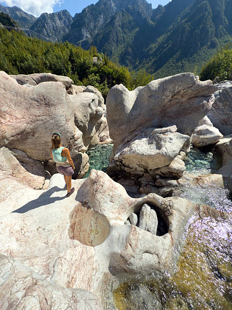 Woman in casual summer clothes standing on a large rock near a clear natural pool. Scenic outdoor setting with rocky terrain and green forested mountains. Nature and adventure concept.
