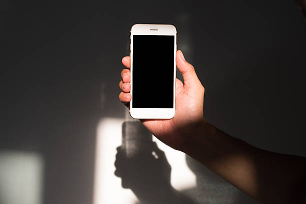 A direct, eye-level shot of a hand holding a white smartphone vertically. The blank black screen is perfectly centered, surrounded by natural sunlight and shadows, making it a perfect mockup for app developers and UI designers.