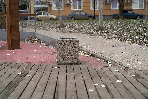 A trash bin sits on wooden planks next to a pathway covered in fallen leaves.