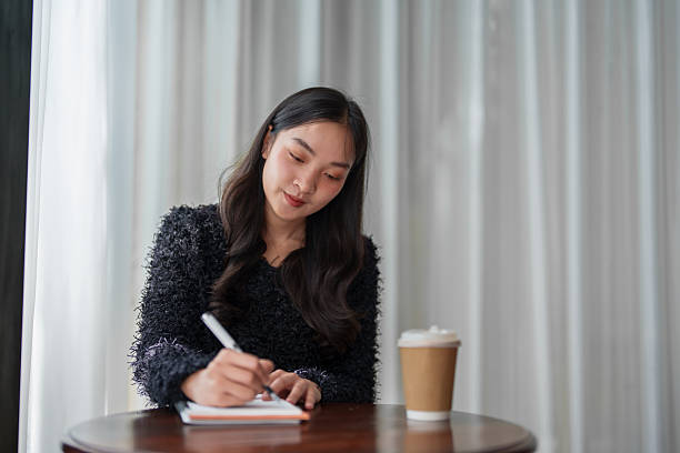 istock Concentrated young asian woman writing notes in notebook while sitting at cafe table with takeaway coffee cup, enjoying quiet atmosphere and natural light 2252142063
