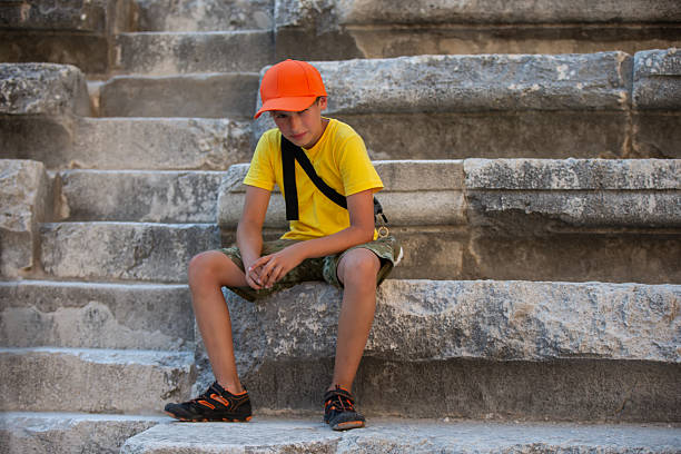 A young boy wearing a yellow shirt and an orange cap takes a break while sitting on the weathered stone steps of ancient ruins. He appears thoughtful as he enjoys the surroundings.