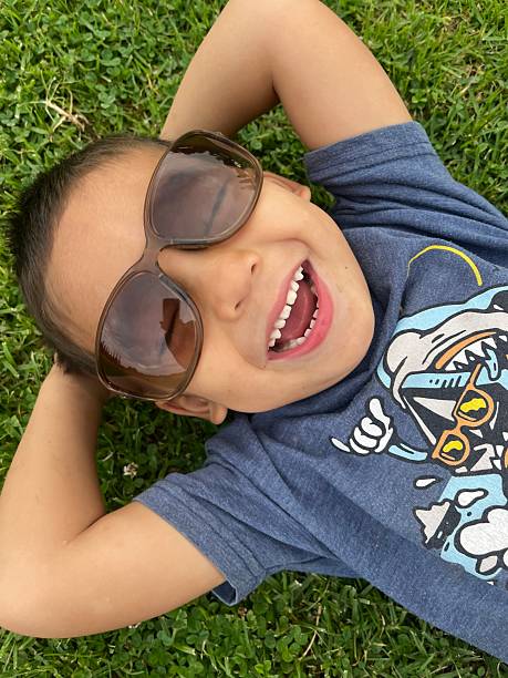 A small child wearing big sunglasses is laying on the grass at the park on a bright sunny day
