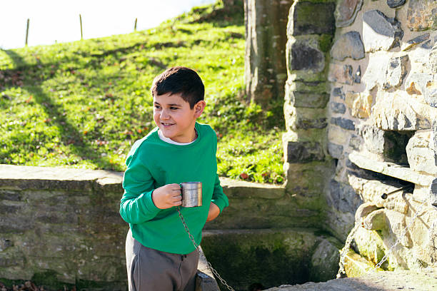 Boy in a green sweatshirt holding a metal cup with a chain, smiling, standing outdoors next to a stone water fountain