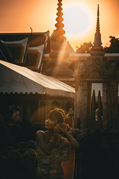 A woman wearing traditional Thai dance attire adjusts her costume at a temple complex in Bangkok, Thailand, during golden hour.