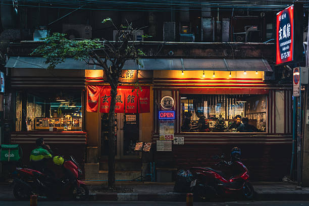 A restaurant exterior is illuminated at night along a city street in Bangkok, Thailand, with diners visible inside and motorbikes parked outside.