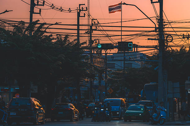 Cars and motorbikes move through a busy intersection at dusk in Bangkok, Thailand, with power lines and a Thai flag visible above the street.