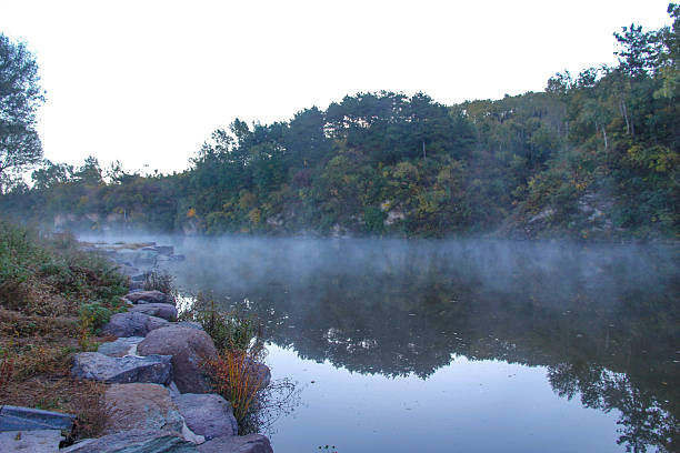 Serene river landscape with mist, reflecting trees and rocks. Forested hills surround the calm water, creating a tranquil natural environment.