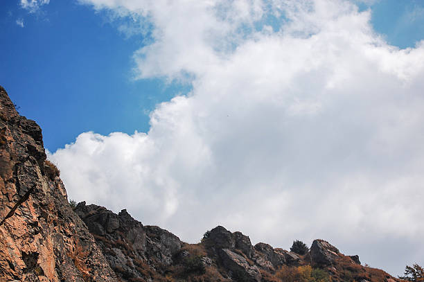 Rocky mountain peak with rugged cliffs under a bright blue sky with fluffy white clouds. Natural outdoor landscape with rocky terrain and open sky.