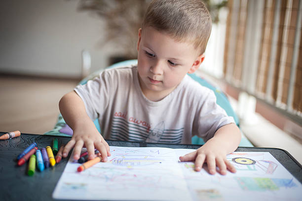 A young boy sits at a table, focused on coloring with crayons. Bright drawings spread out before him, capturing his imagination as he creates art in a cozy indoor space.